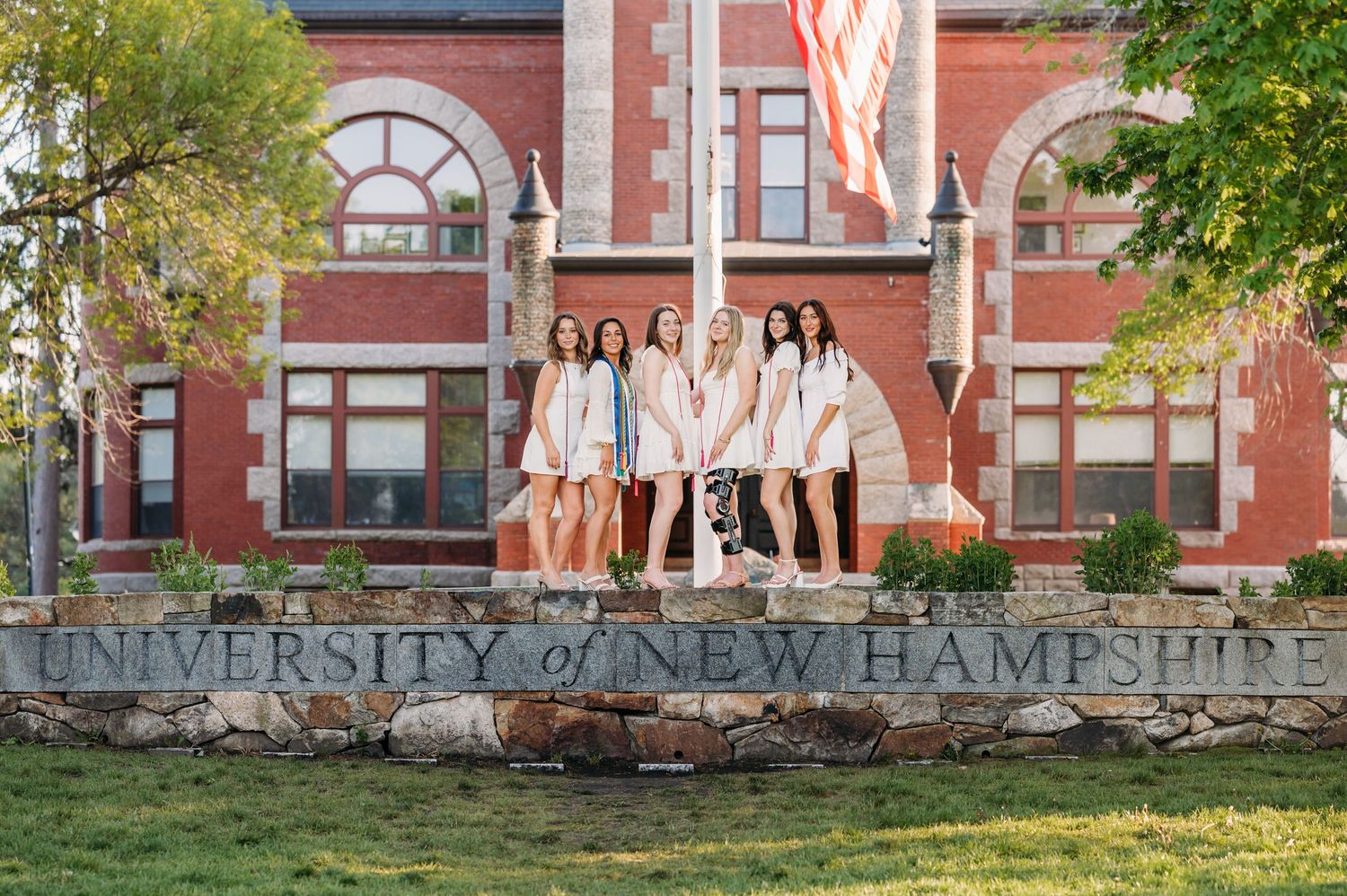 College students sitting on stone sign at University of New Hampshire campus with red brick building in background.