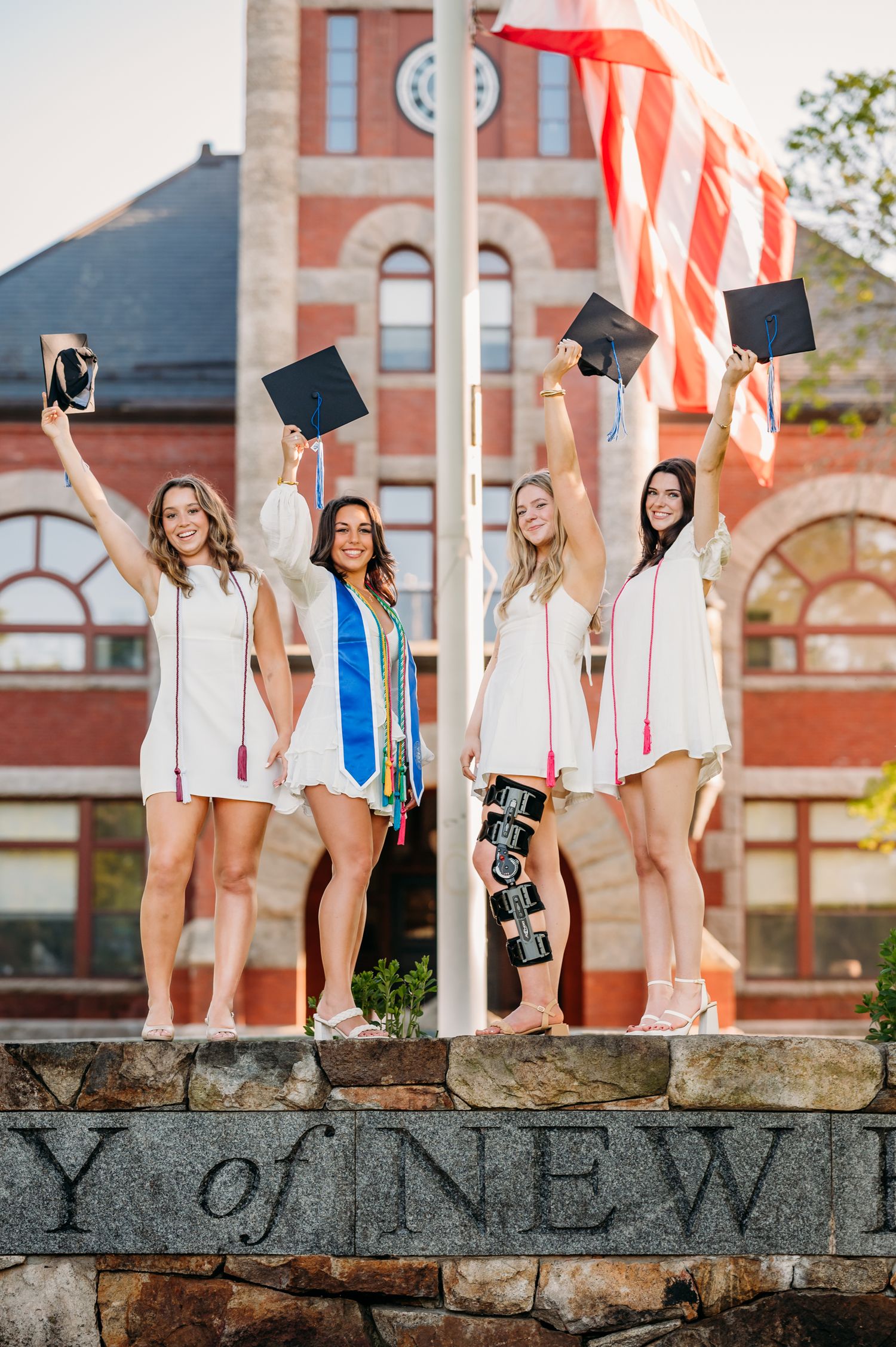 College graduates in white dresses celebrate with caps and diplomas in front of historic brick building with American flag.
