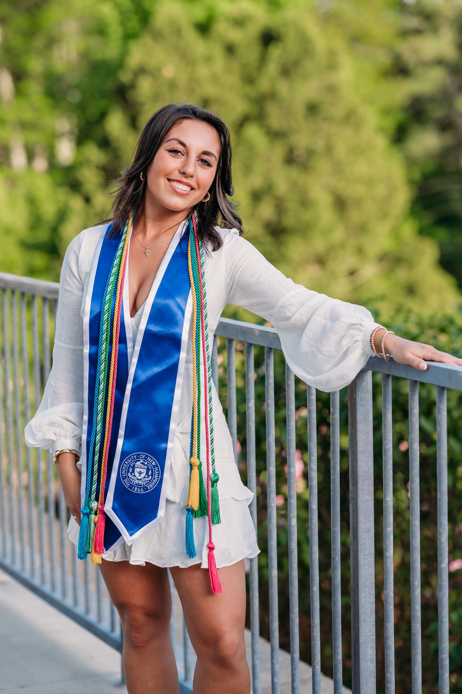 Graduate in white dress and blue graduation stole poses on bridge during outdoor photo session.
