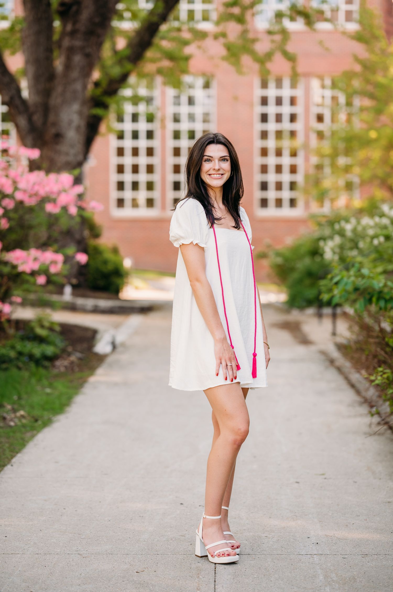 A person in a white dress with puff sleeves stands outdoors on a pathway with pink flowers and buildings in the background.