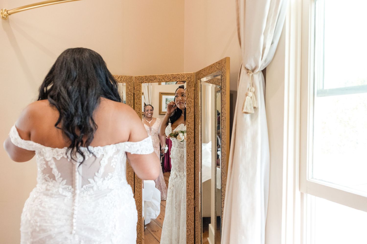 A person in a white lace wedding dress stands in front of an ornate mirror while natural light streams through a nearby window.