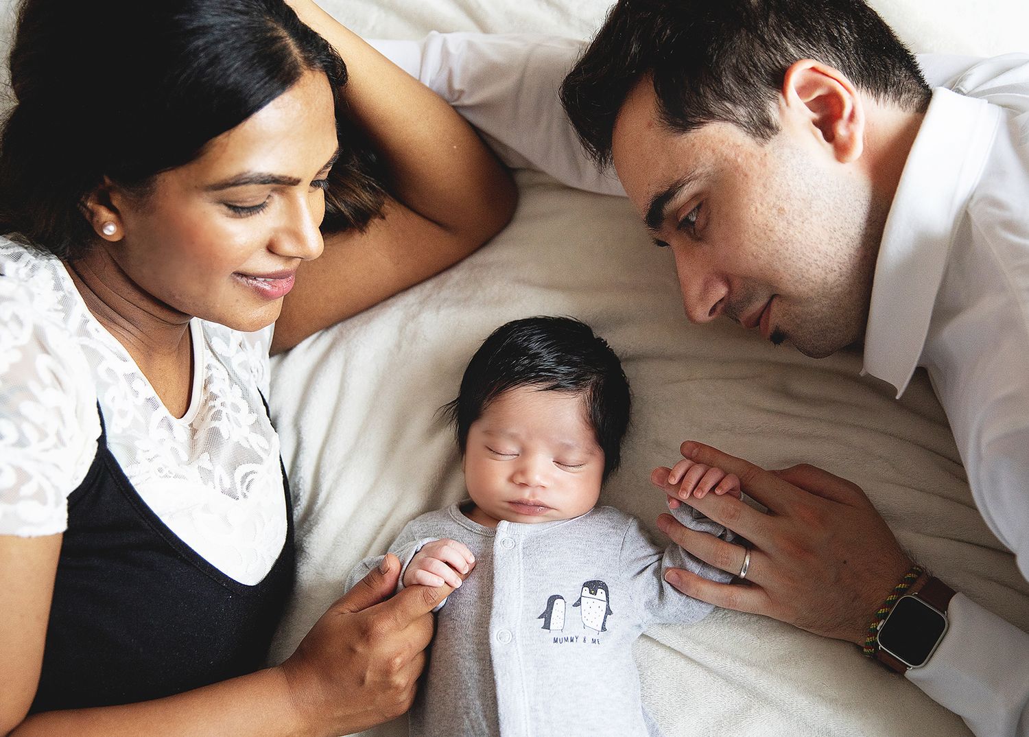 Couple and their newborn baby share a tender moment while laying together on white bedding. During a home newborn session in London