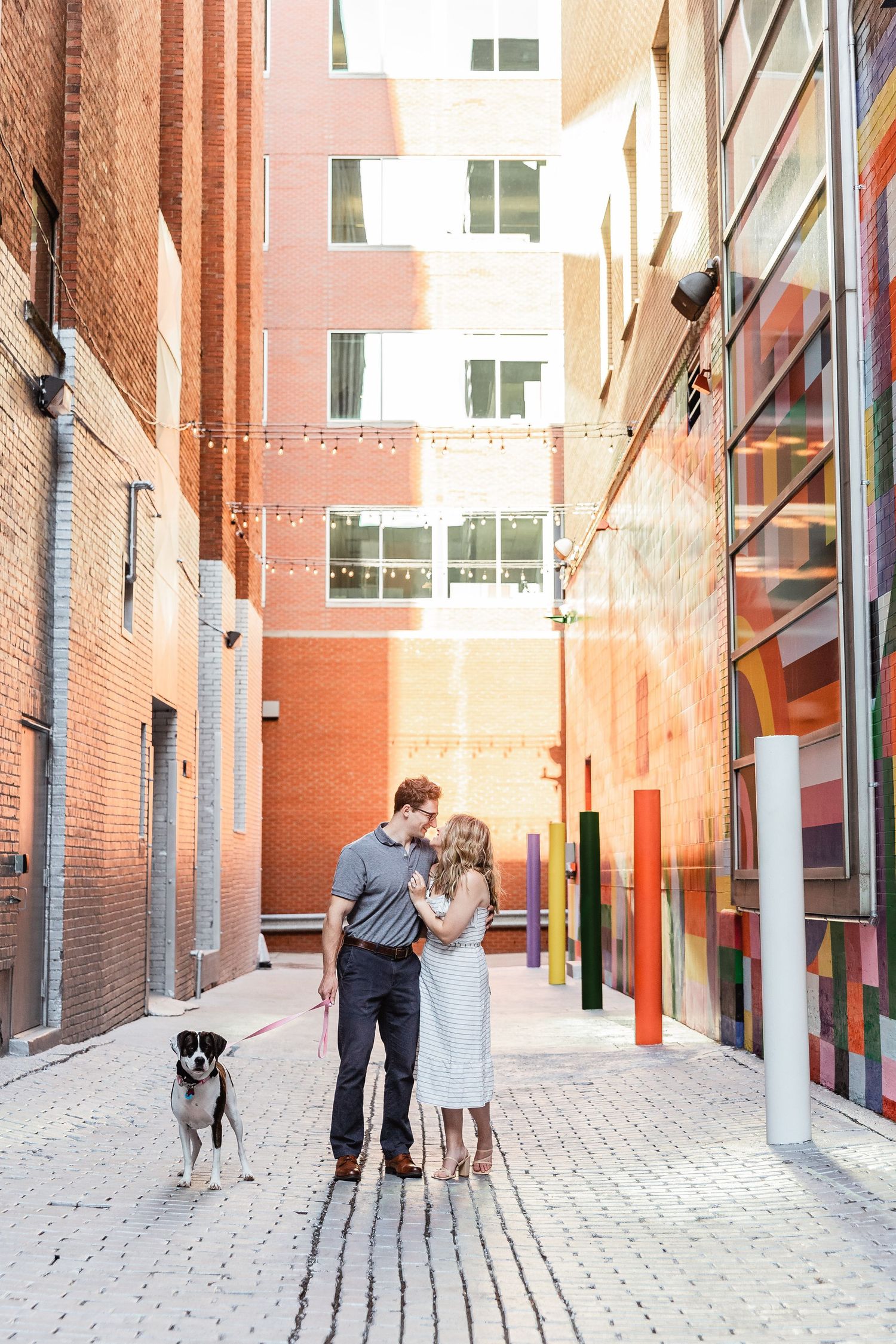A couple kneels with their fluffy gray and white dog on a rooftop terrace overlooking a cityscape at sunset.