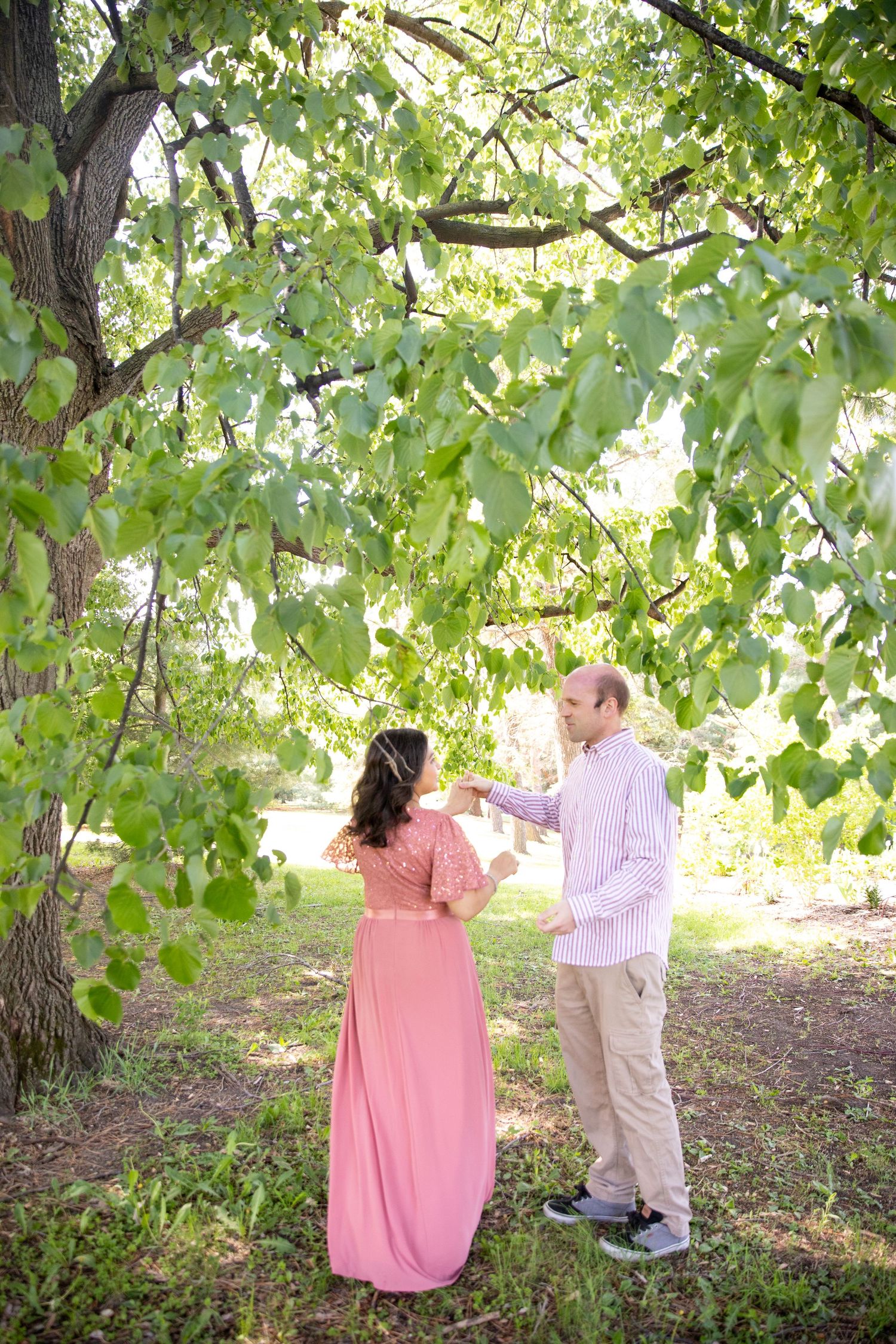 A couple shares a tender moment under dappled sunlight filtering through green tree leaves in a natural outdoor setting.
