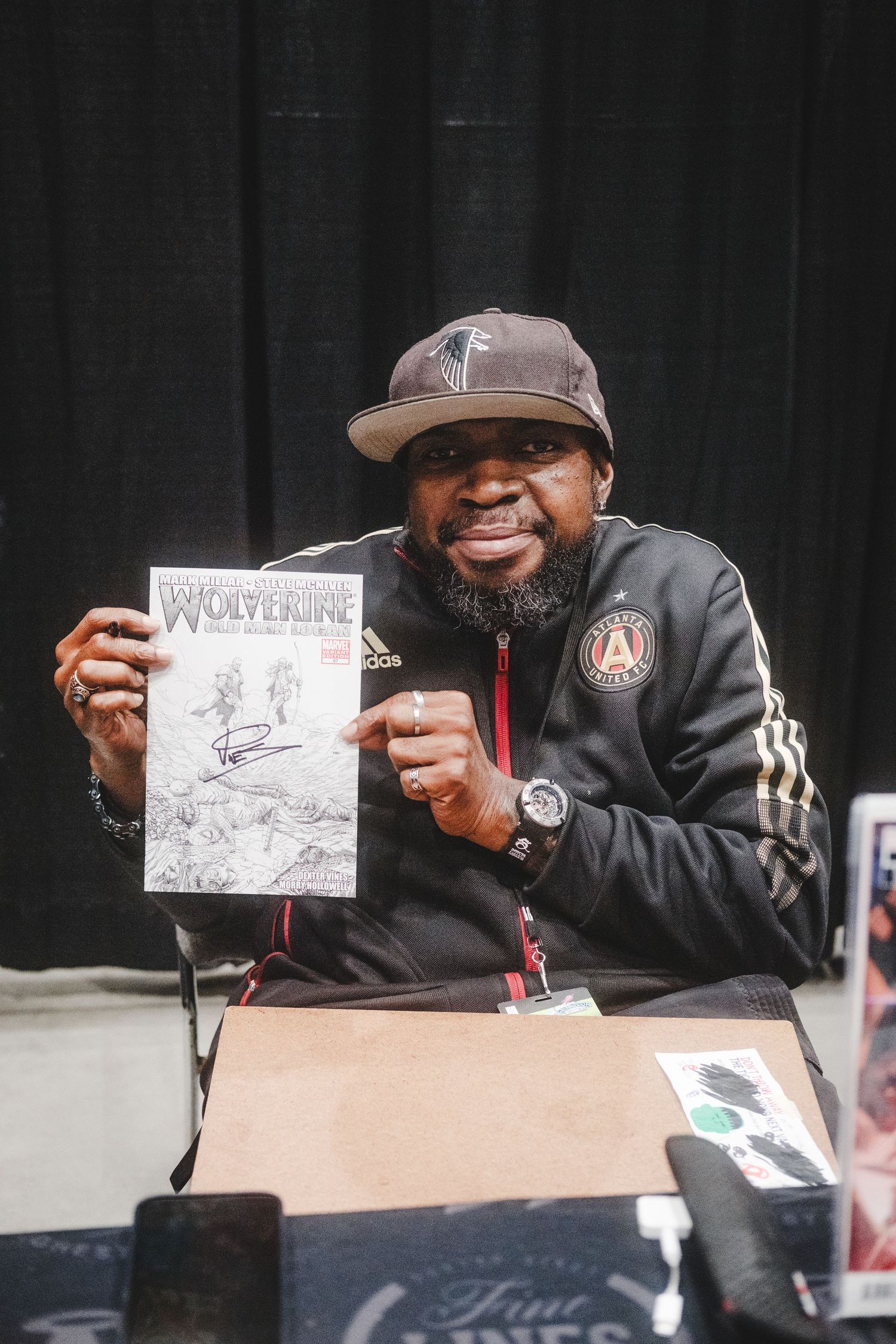 Person in black Adidas jacket and baseball cap smiles while holding up artwork at signing event.
