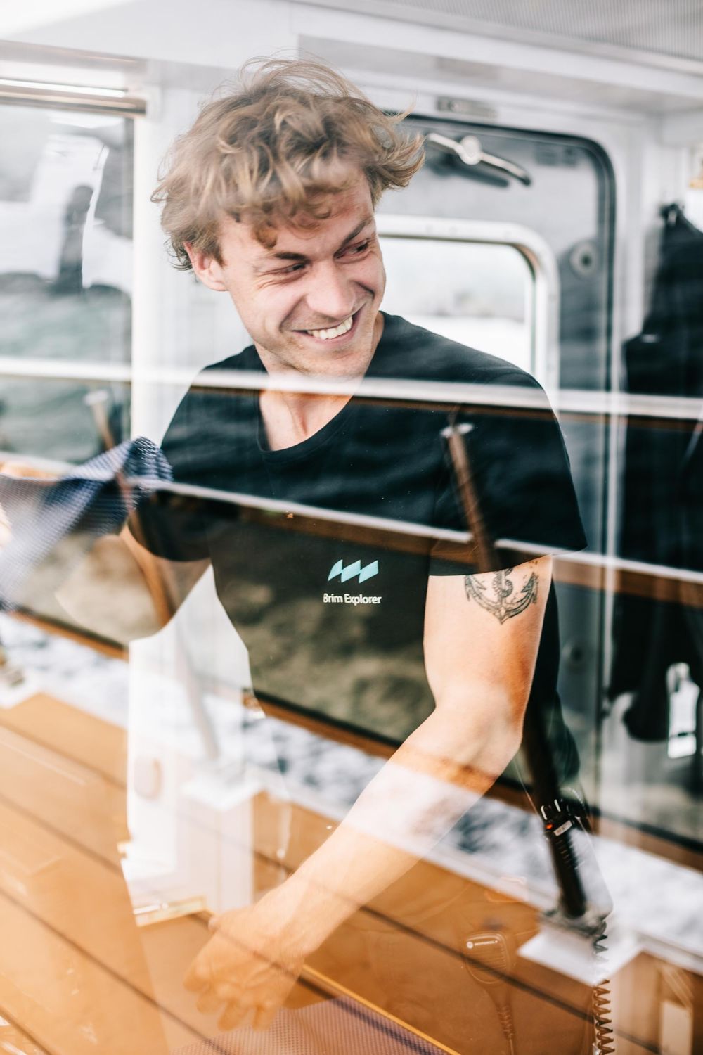 Person in black shirt smiling through window reflection with bright natural lighting.