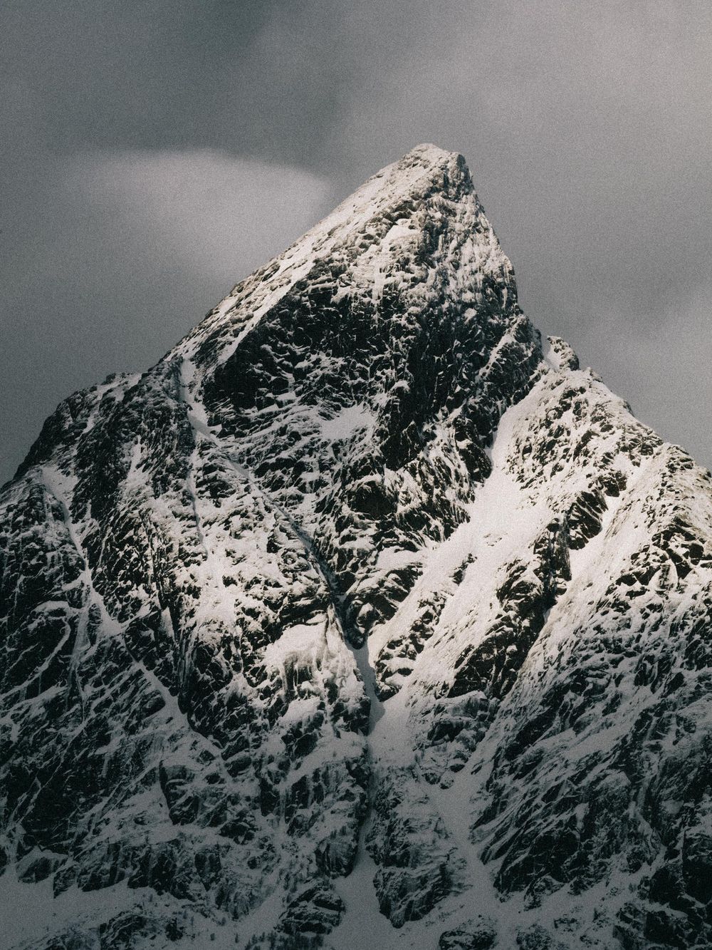 A dramatic snow-covered mountain peak rises against a moody gray sky in stark black and white contrast.