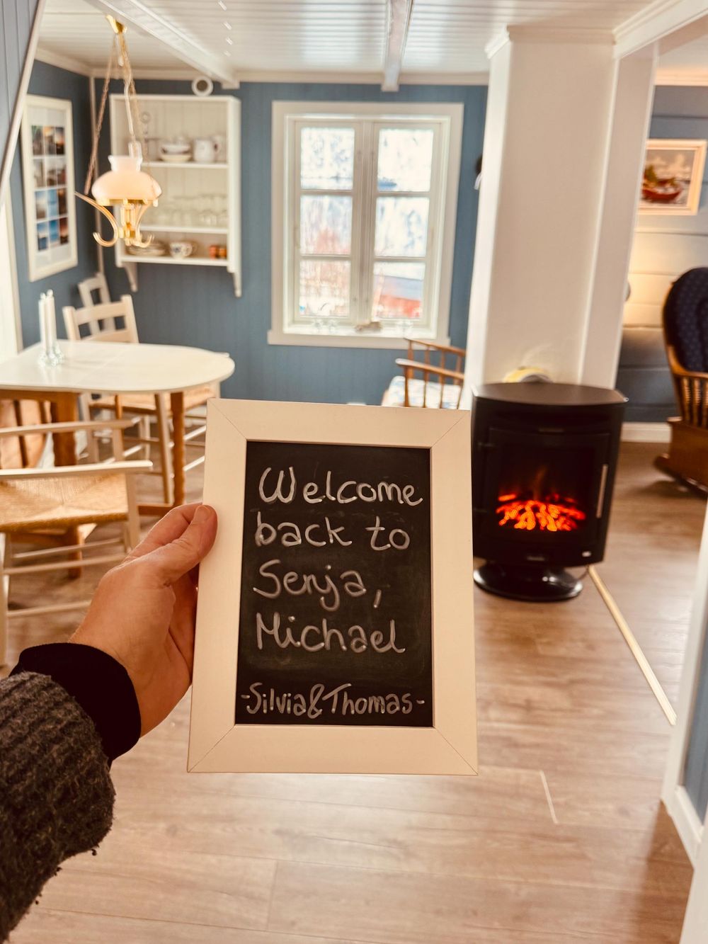 Cozy dining room interior with blue walls, wooden furniture, and a hand holding a welcome chalkboard sign near a wood stove.