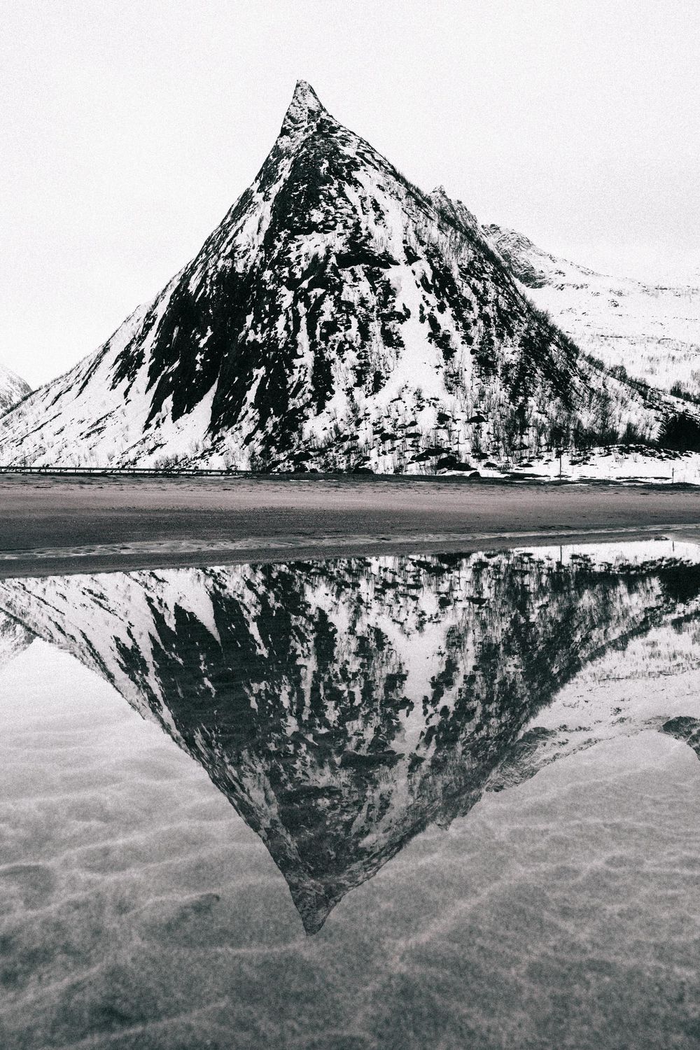 A dramatic black and white mountain peak perfectly reflected in calm waters of a Nordic fjord in winter.