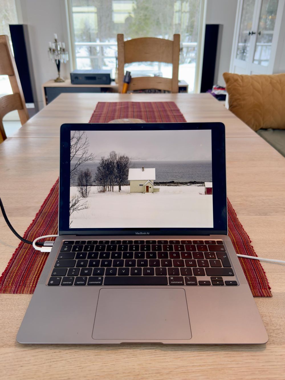 MacBook laptop on wooden table displays snowy winter scene with small white house and bare trees on screen.