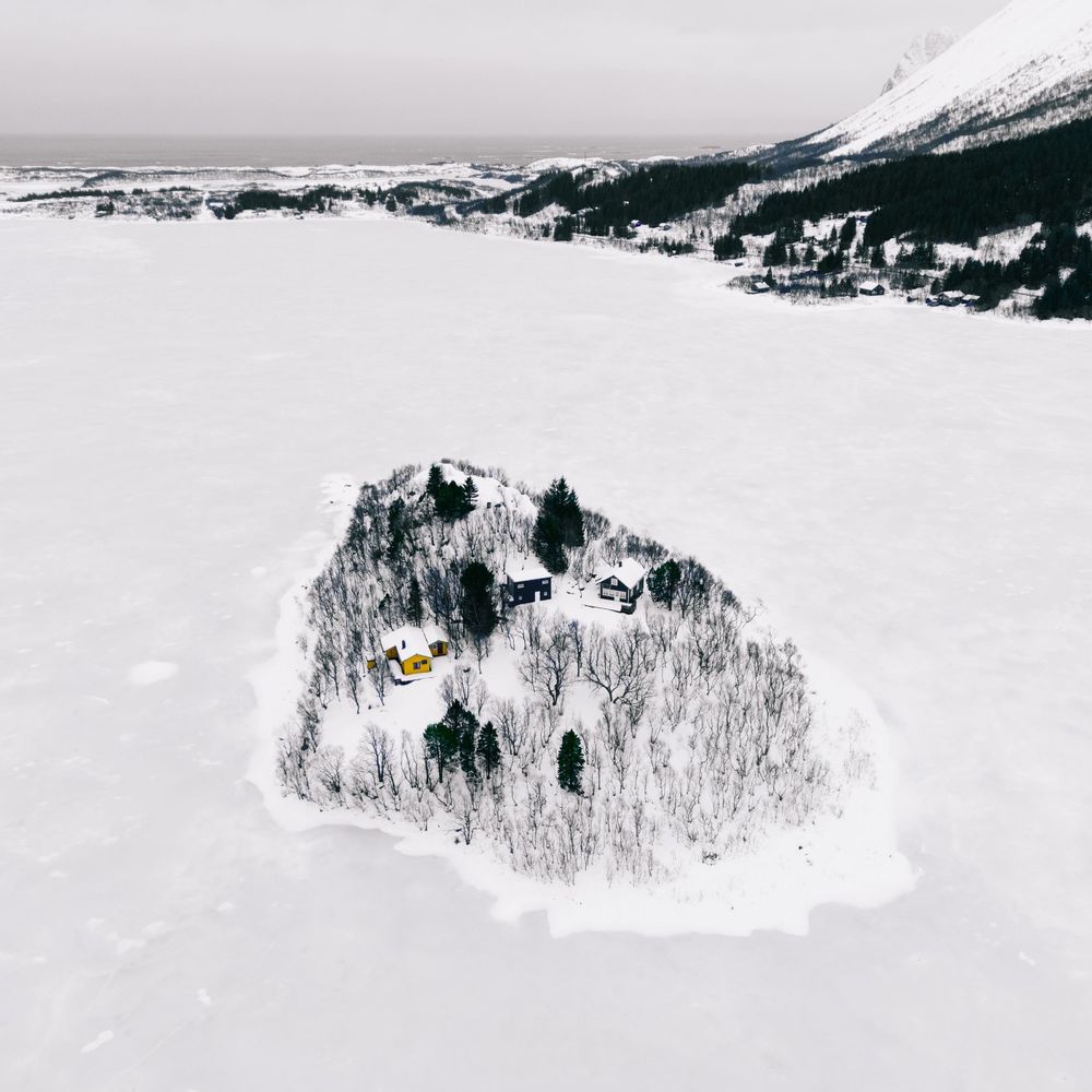 Aerial winter view of a small snow-covered island with trees and buildings surrounded by frozen lake landscape.