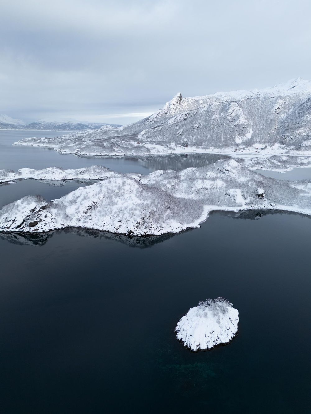 Aerial view of snow-covered mountains surrounding dark waters with a small snow-capped island in Norwegian fjords.