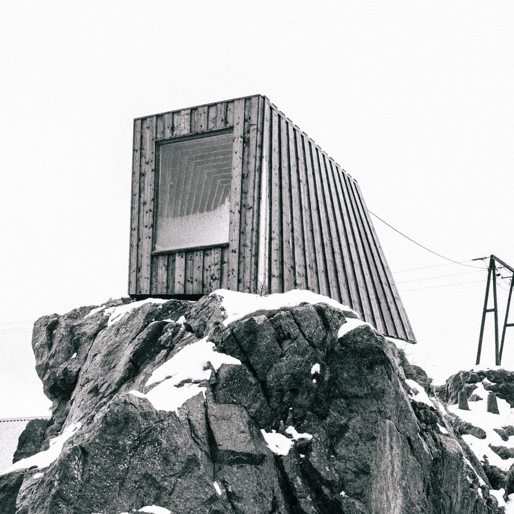 A modern angular wooden shelter perched on snowy rocks with power lines visible in the background.