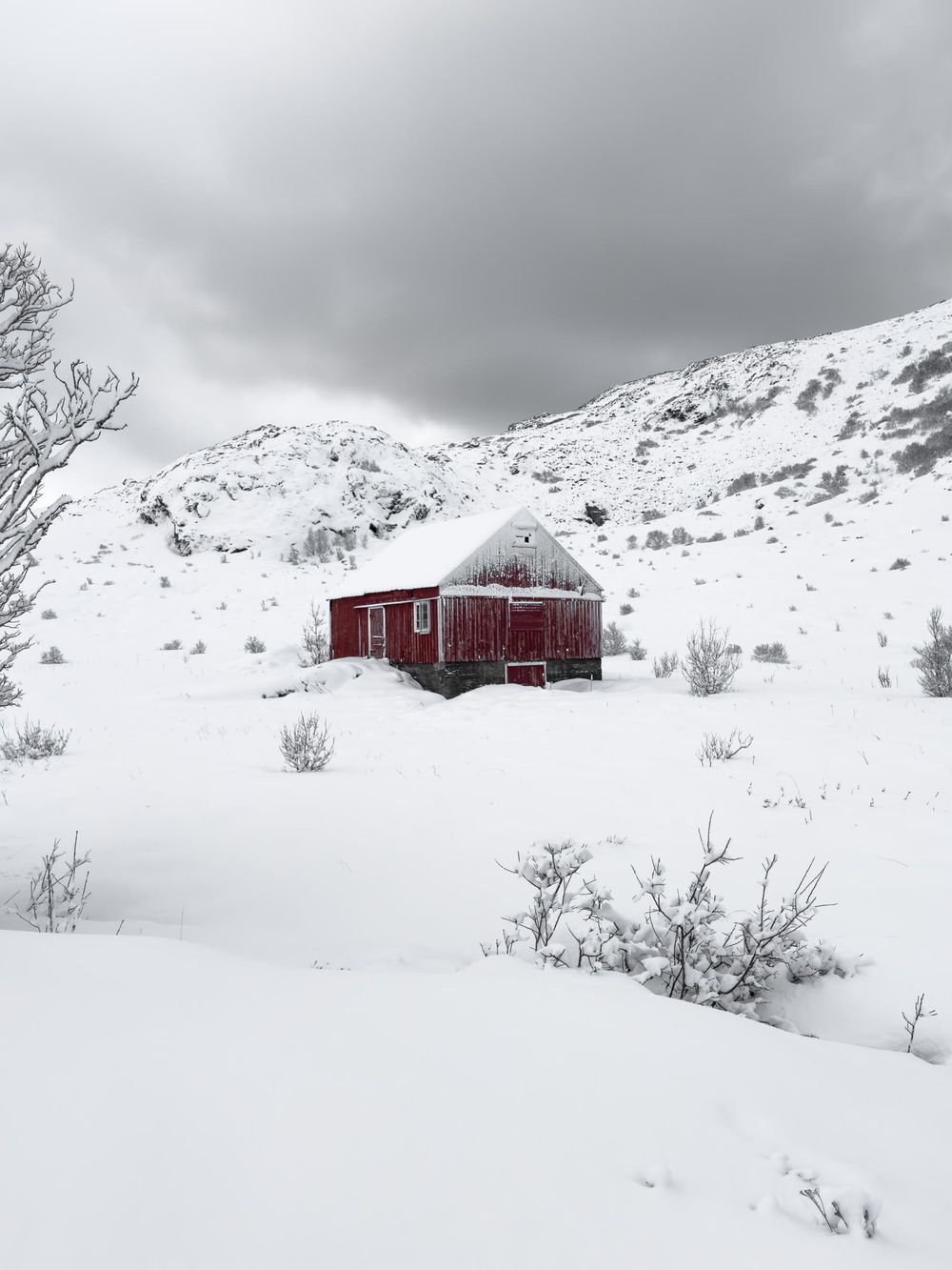 A red wooden cabin stands isolated in a snowy mountain landscape under gray winter skies.