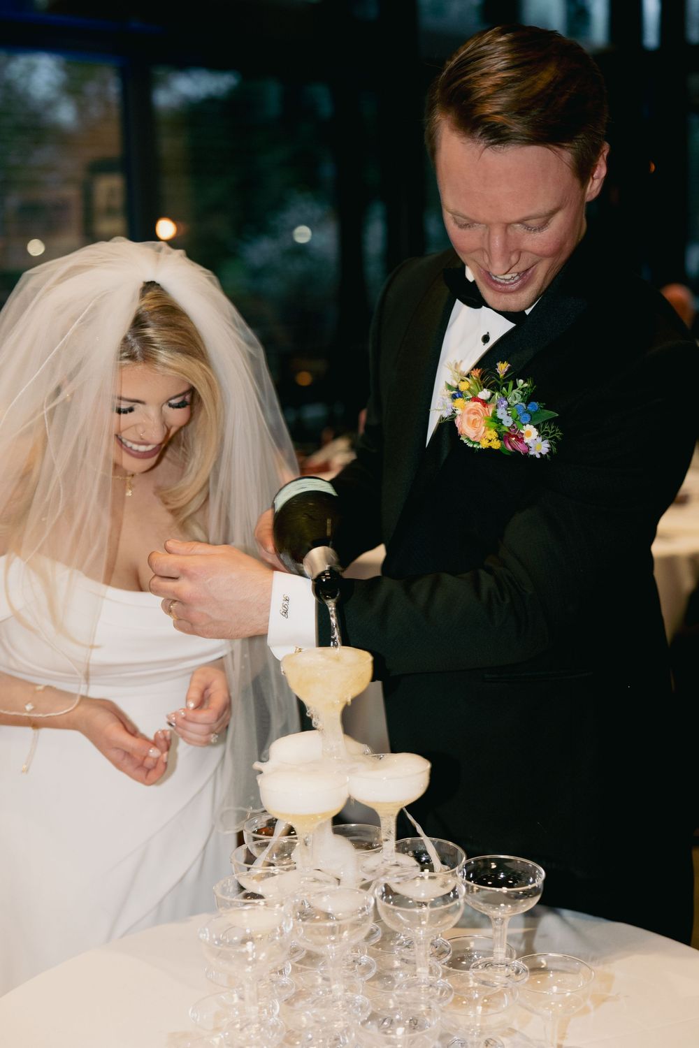 A bride and groom pour champagne into a tower of crystal glasses at their wedding reception.