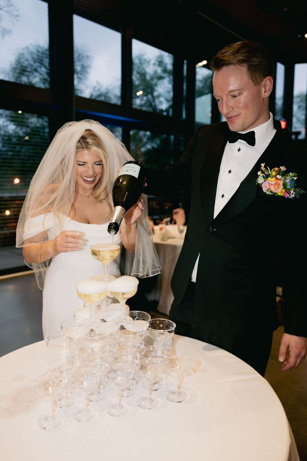 A wedding couple pouring celebratory champagne into a tower of stacked glasses.