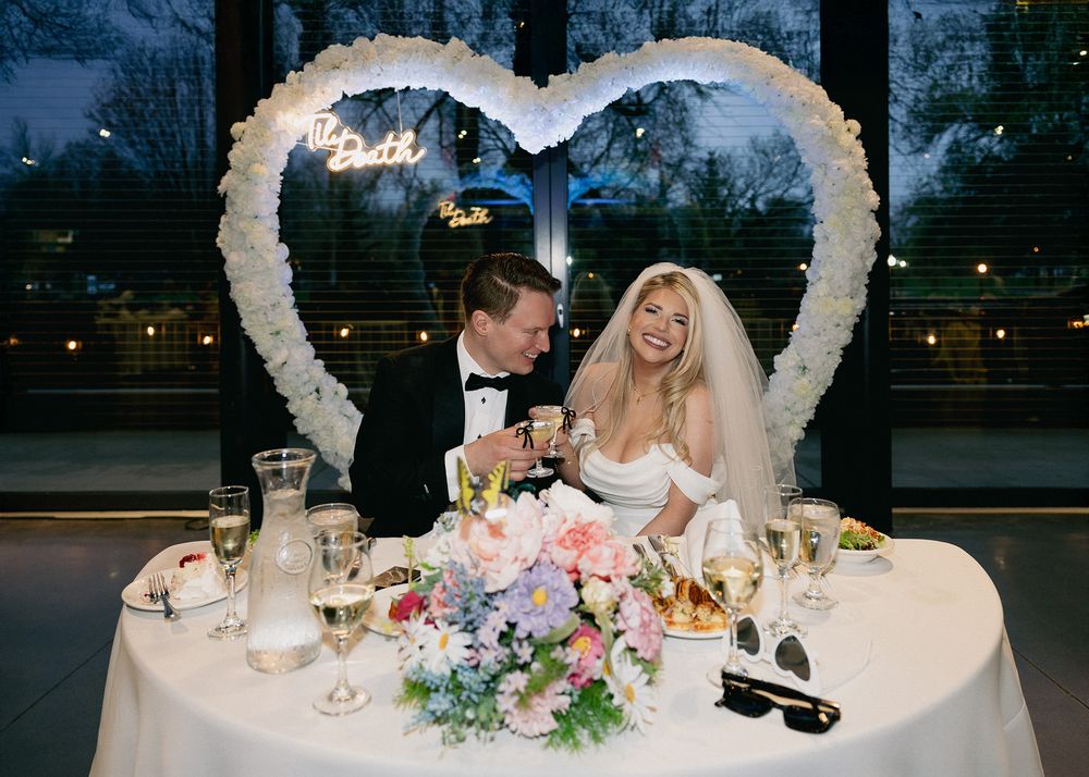 A wedding reception table decorated with a large heart backdrop, floral centerpiece and string lights at an evening venue.