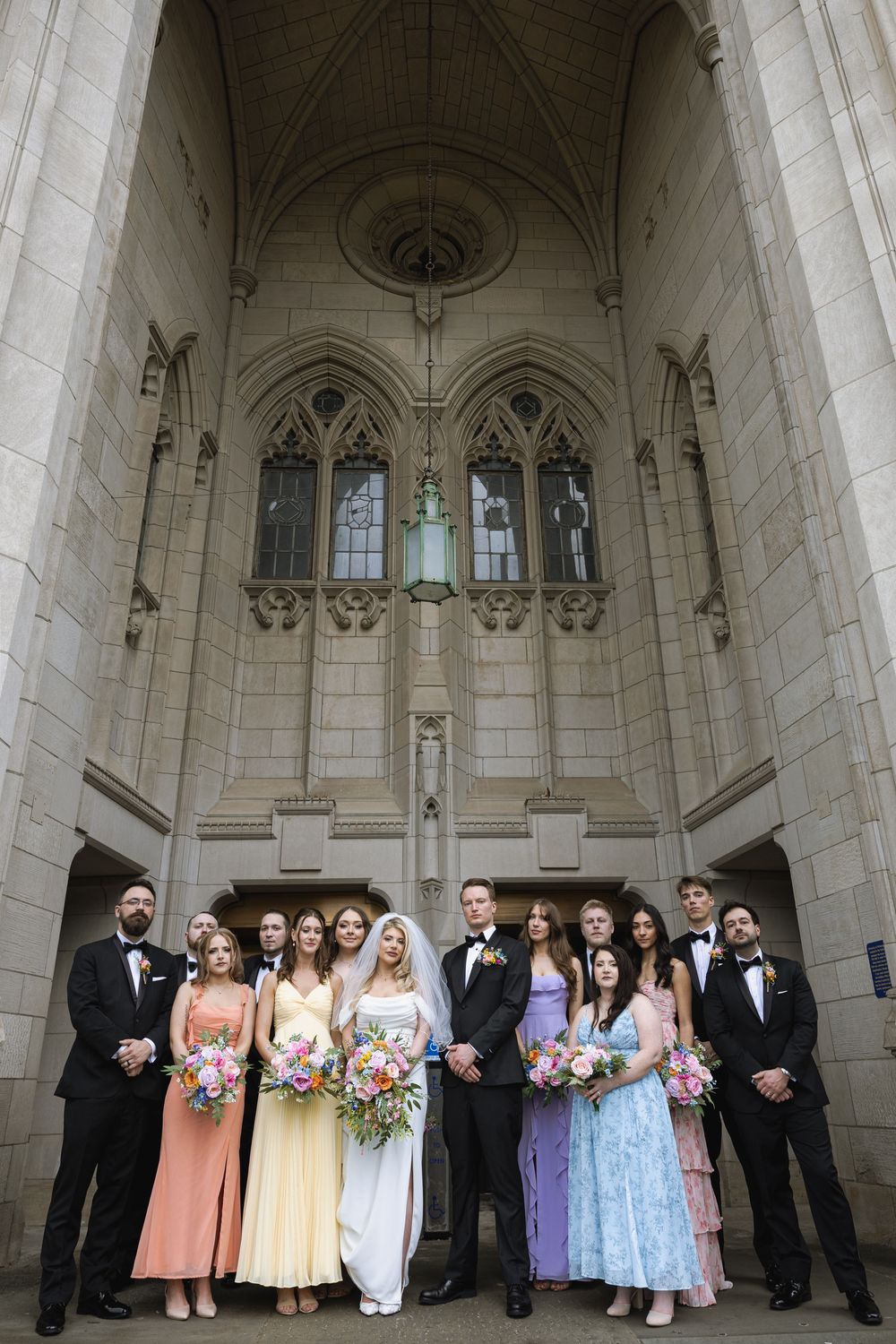 Wedding party in formal attire posed beneath Gothic church archway with stained glass windows and vaulted ceiling.