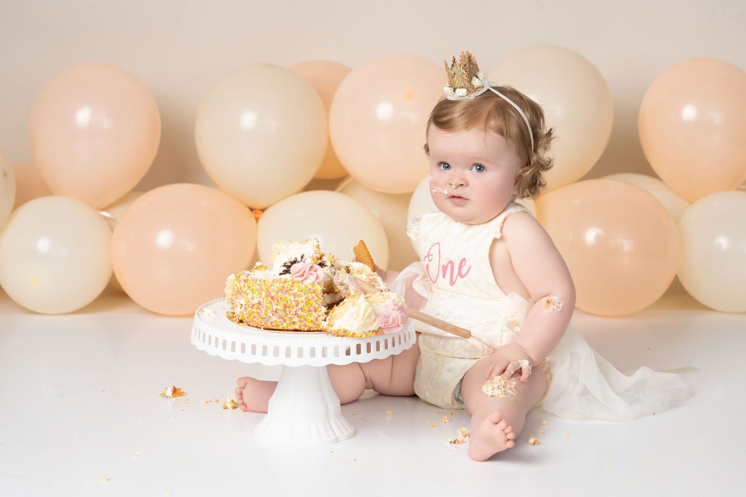Baby in pink outfit celebrates first birthday with cake smash photo session against peach and white balloon backdrop.
