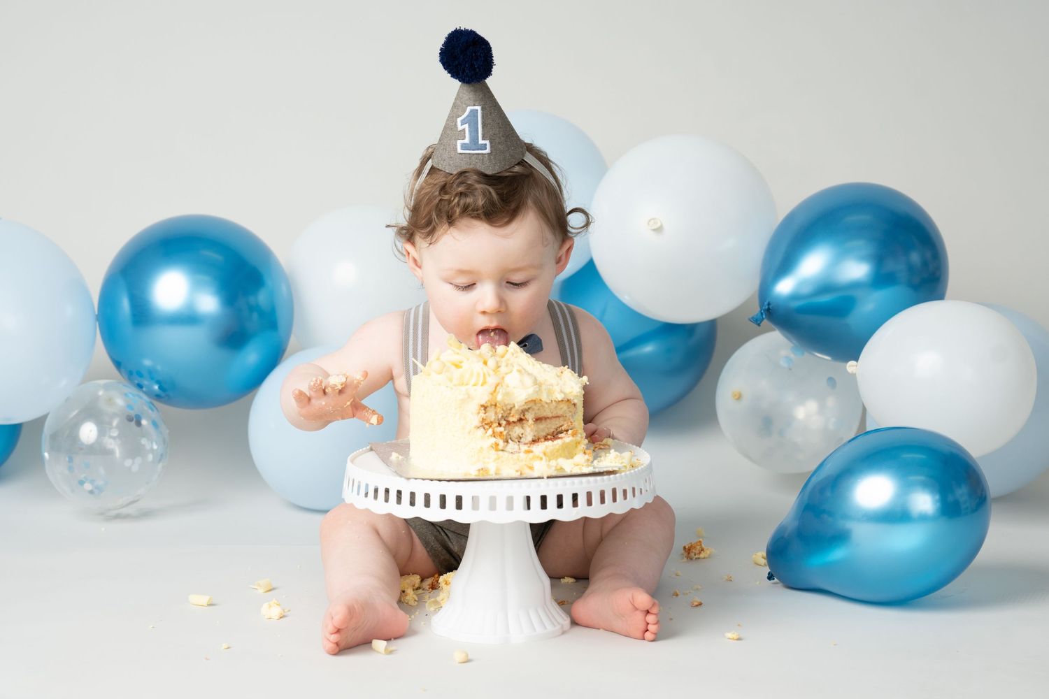 Little one in party hat tastes their first birthday cake on white pedestal surrounded by blue and white balloons.