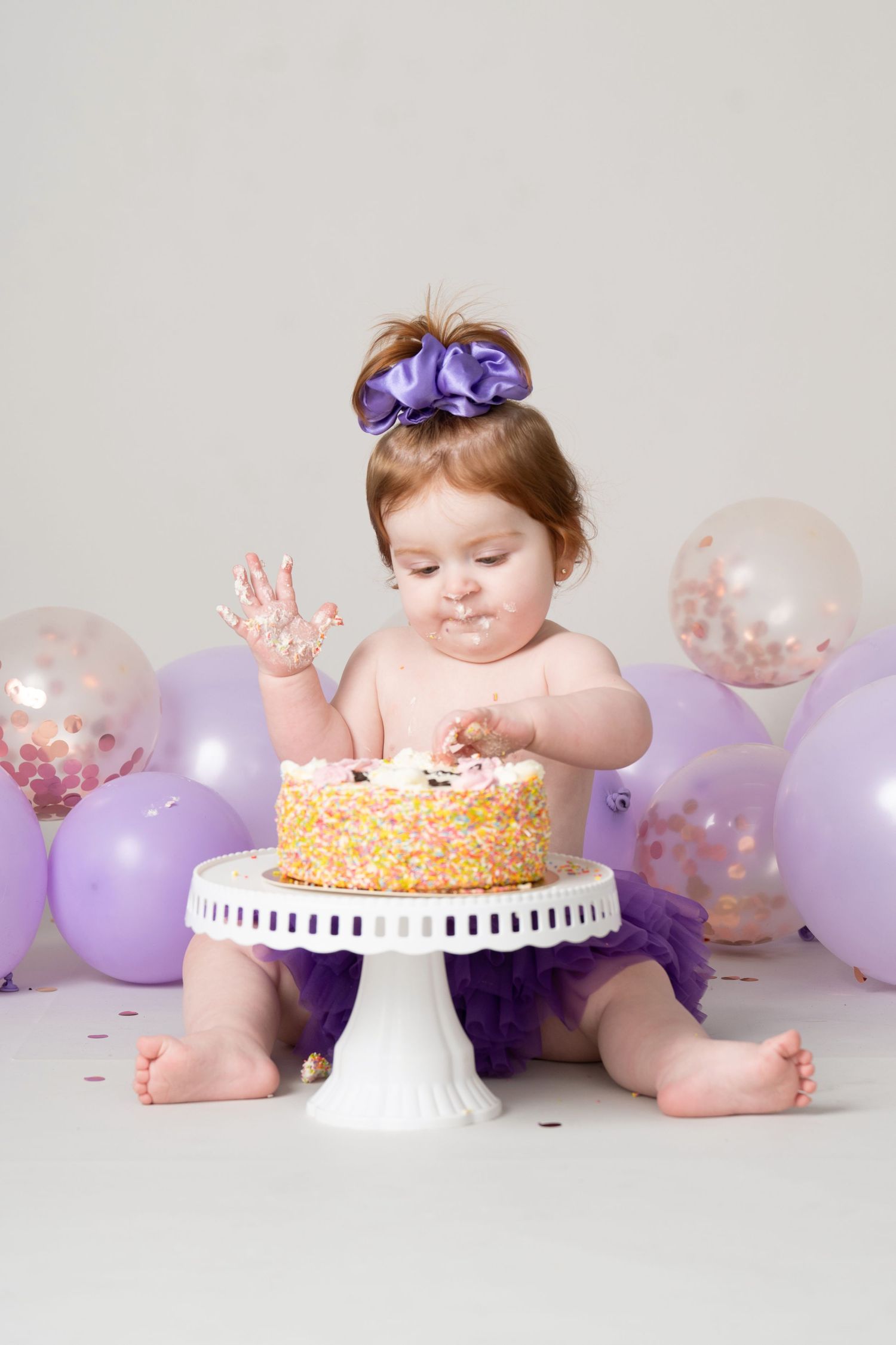 Baby in pink dress with purple bow explores first birthday cake on white stand surrounded by purple and confetti balloons.