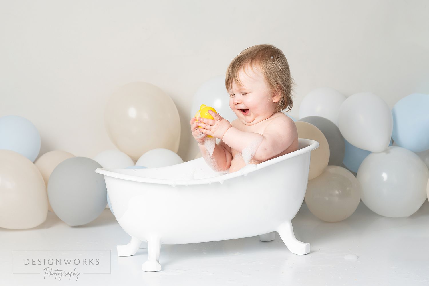 Baby sits in miniature white bathtub surrounded by pastel balloons while playing with yellow toy duck.