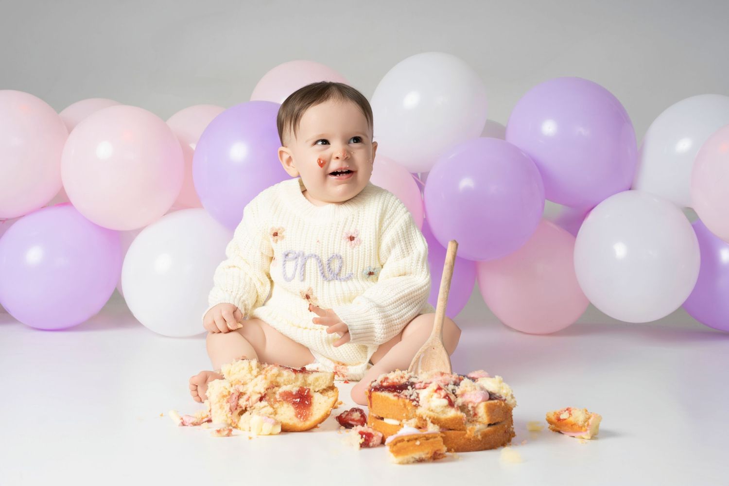 A smiling baby seated with cake smash setup featuring pink, purple and white balloons in the background.