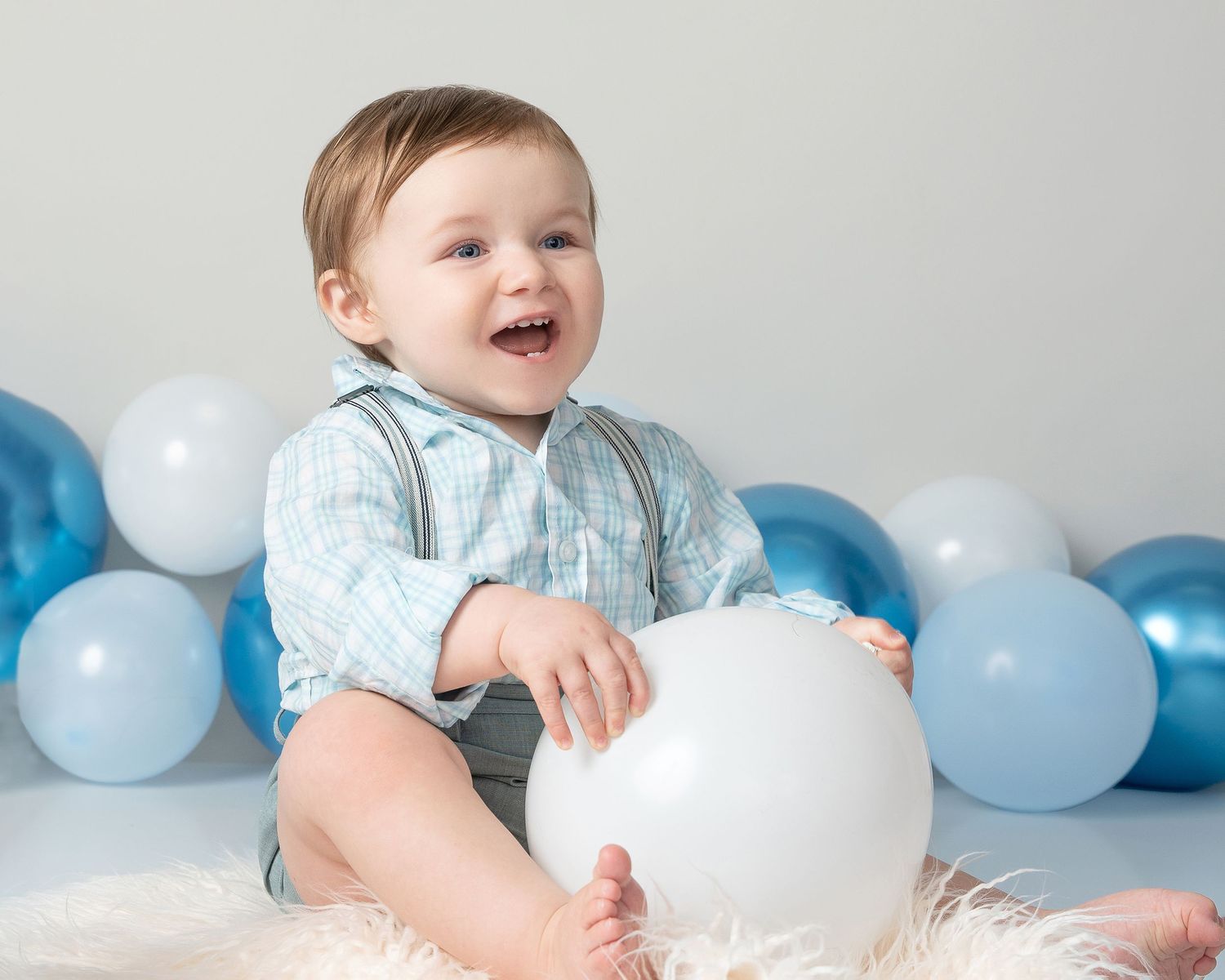 A smiling baby in a light blue outfit sits on a fluffy rug surrounded by blue and white balloons.