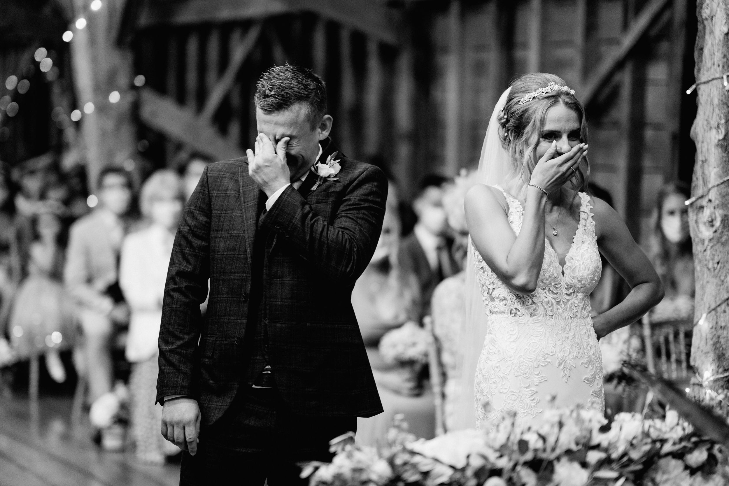 Emotional reactions during a wedding ceremony in a rustic barn venue with white string lights and floral decor.