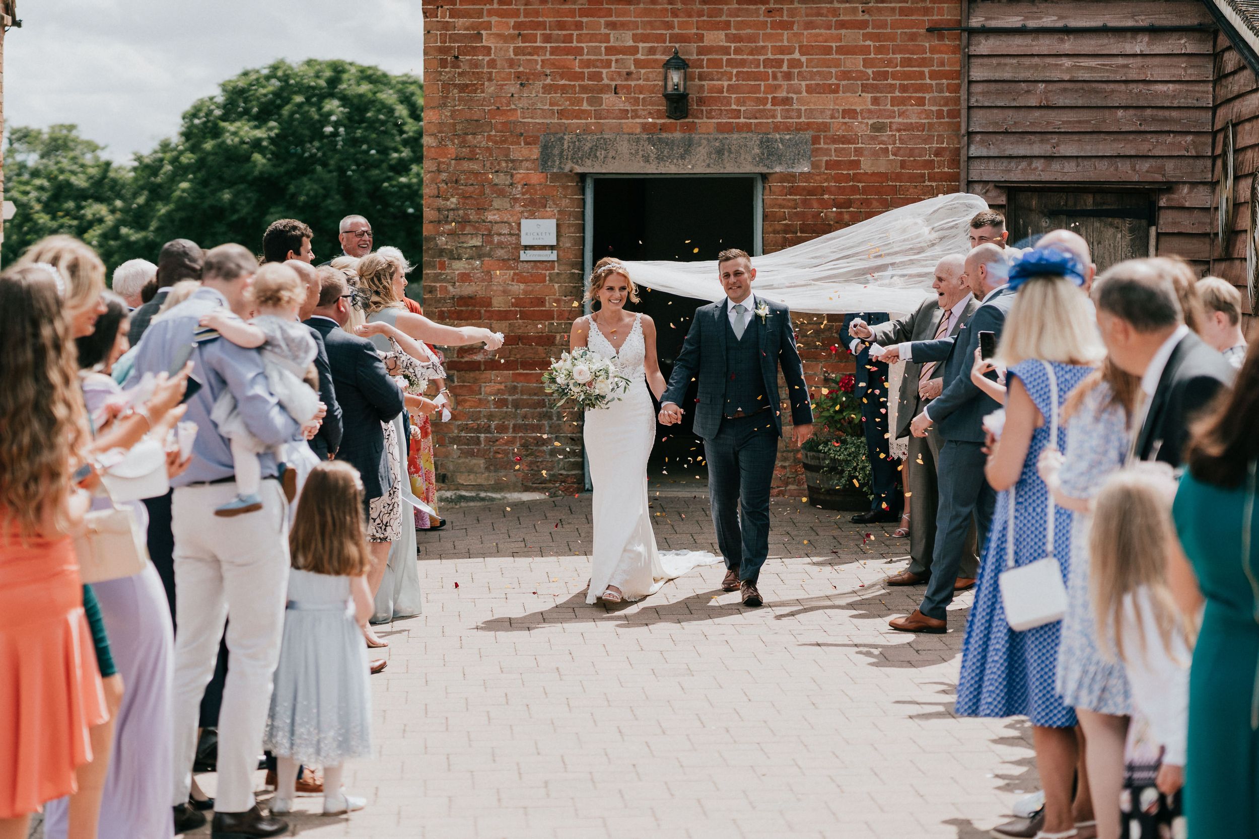 Wedding guests form an aisle outside a brick building to celebrate the newlyweds' exit outside Rickety barn, Bassmead.
