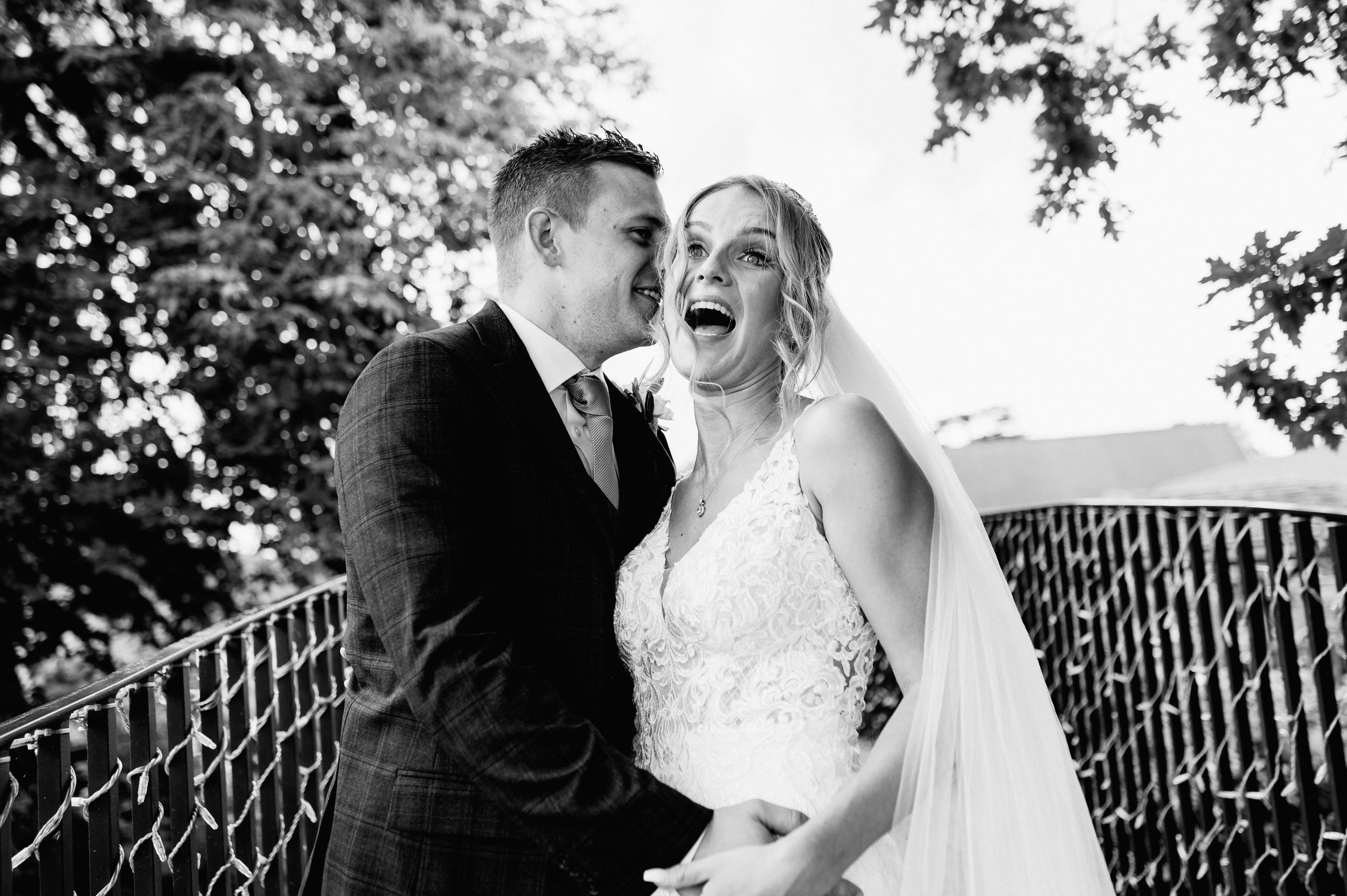 Black and white wedding portrait capturing a candid moment of laughter on an outdoor bridge at Bassmead Manor.