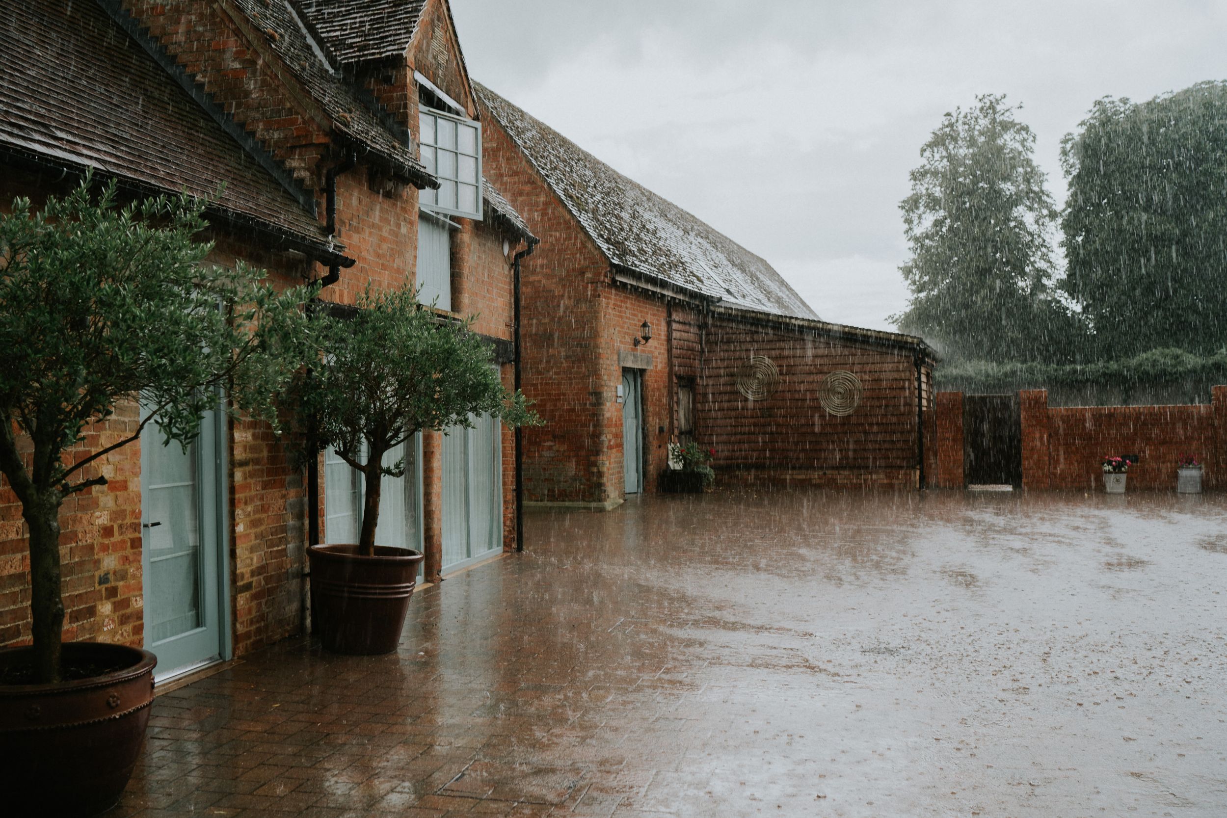 Rain falls heavily on a rustic brick building at Bassmead Manor with blue shutters and potted plants.