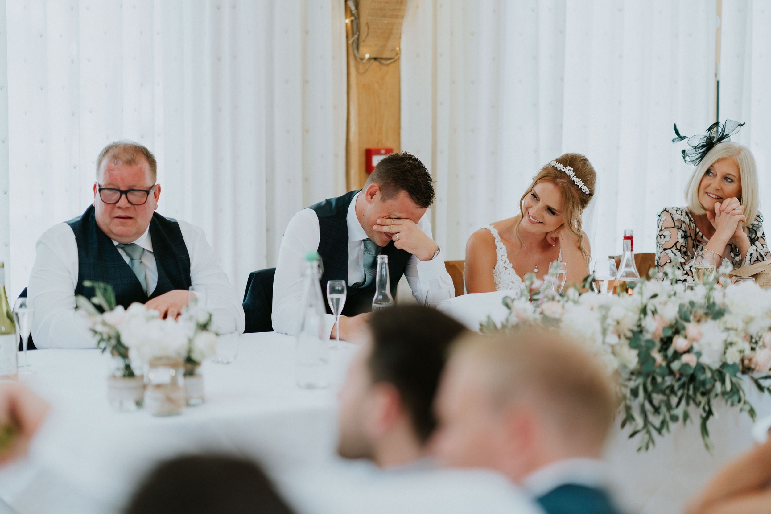 Wedding reception guests seated at tables with floral centerpieces during dinner speeches.
