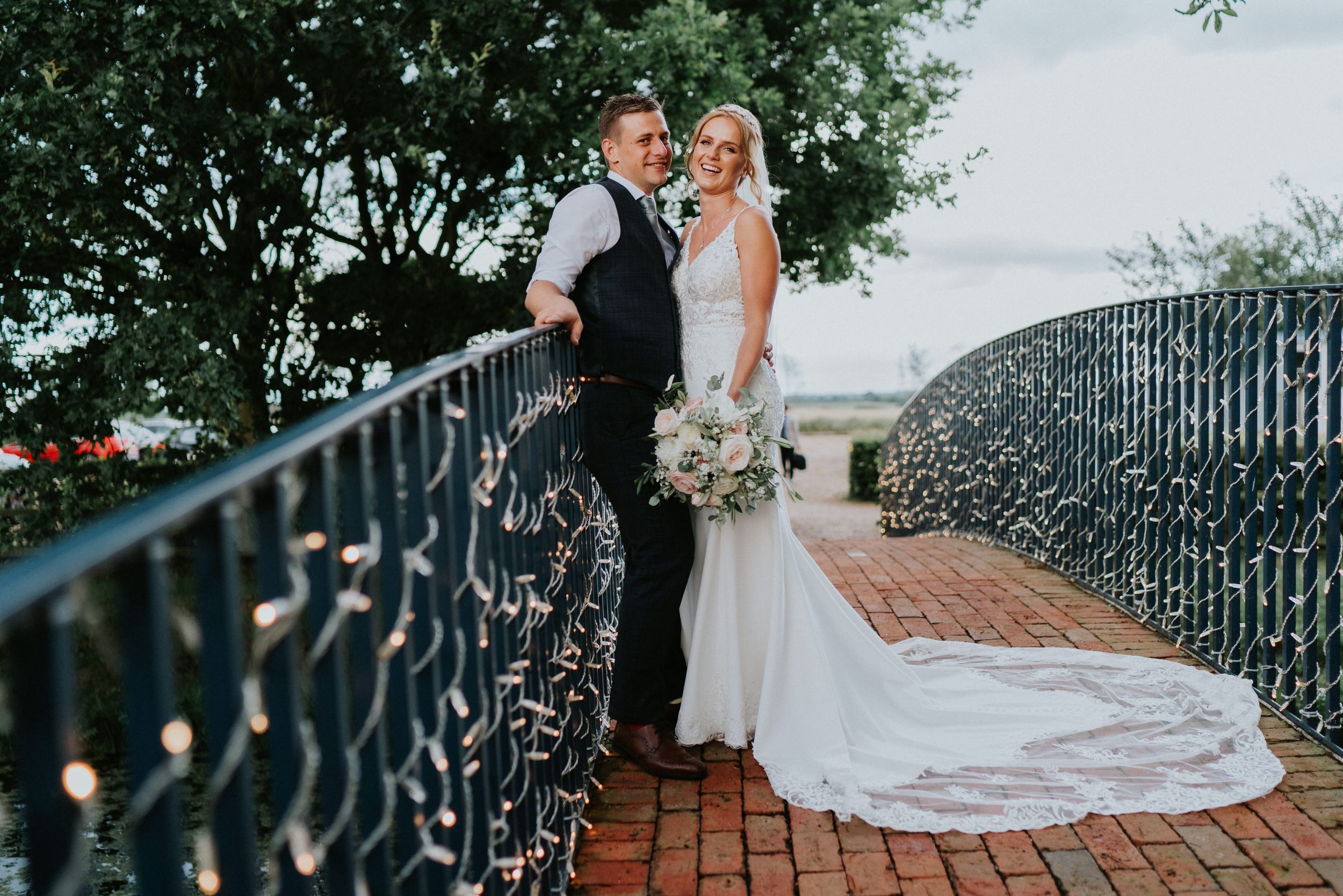 Wedding couple standing on brick terrace with ornate iron railing during sunset.