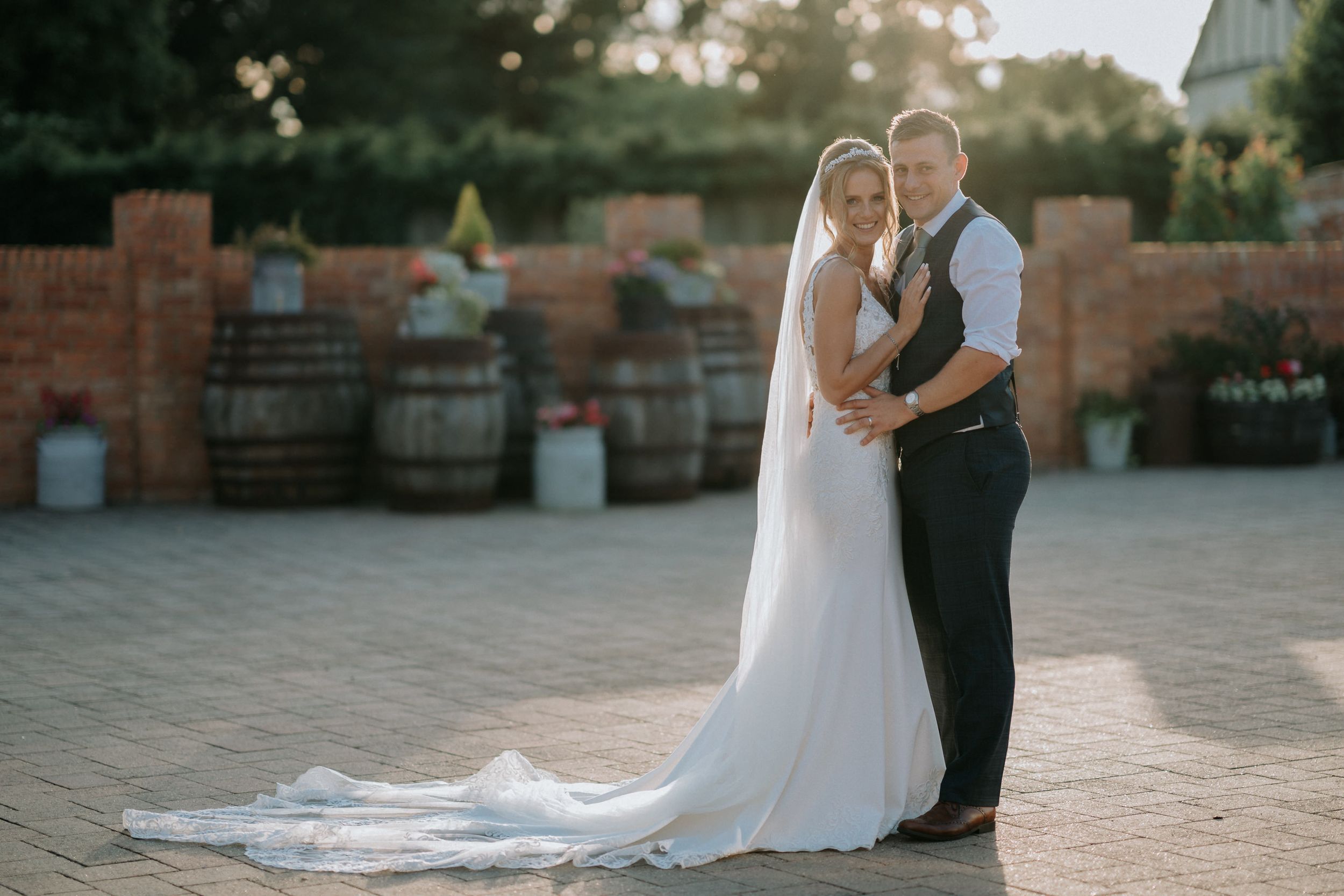 Sunset wedding portrait at Bassmead Manor with couple against brick wall and wine barrels.