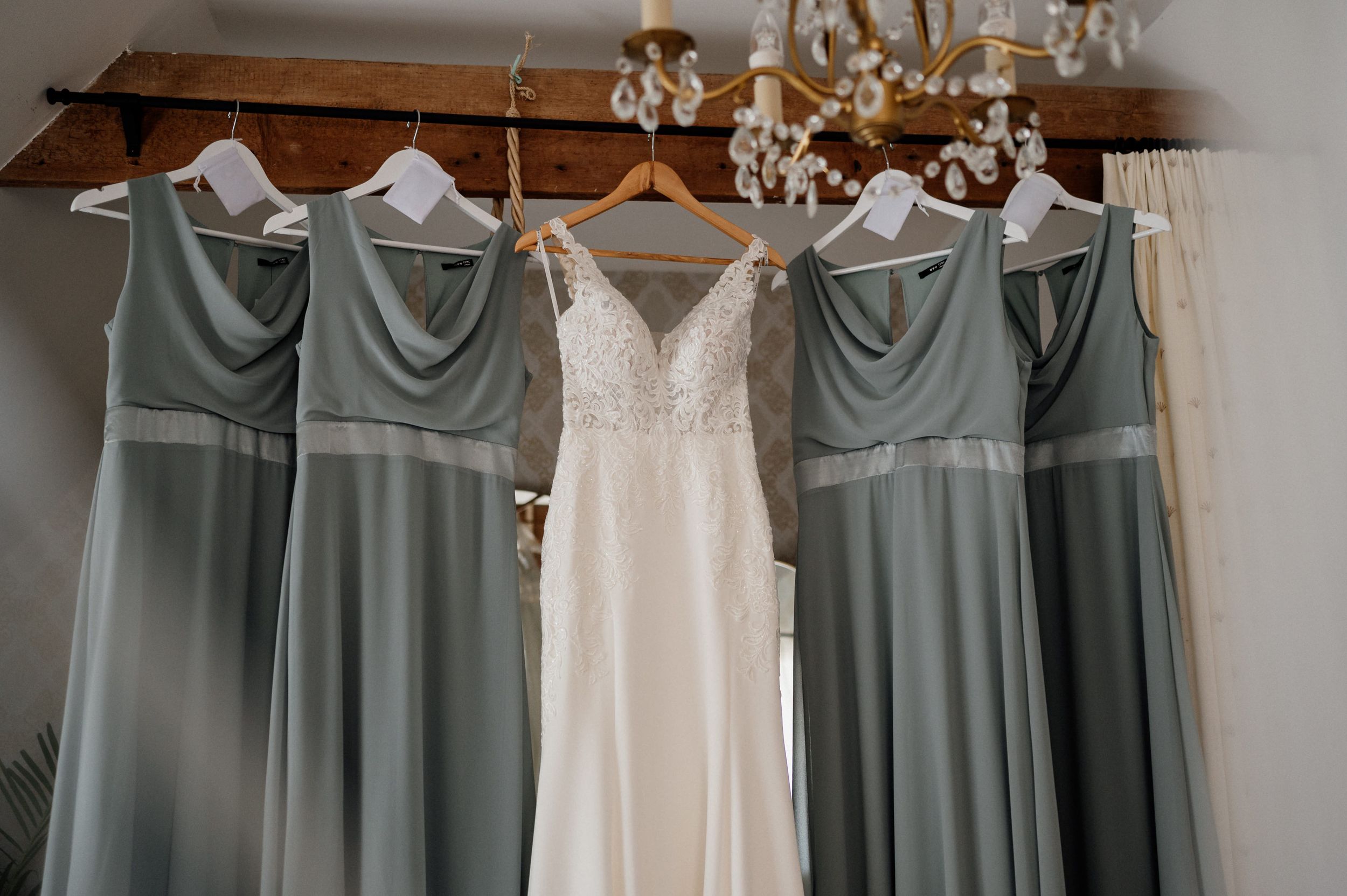 Sage green bridesmaid dresses and white wedding gown hanging on wooden rack beneath crystal chandelier.