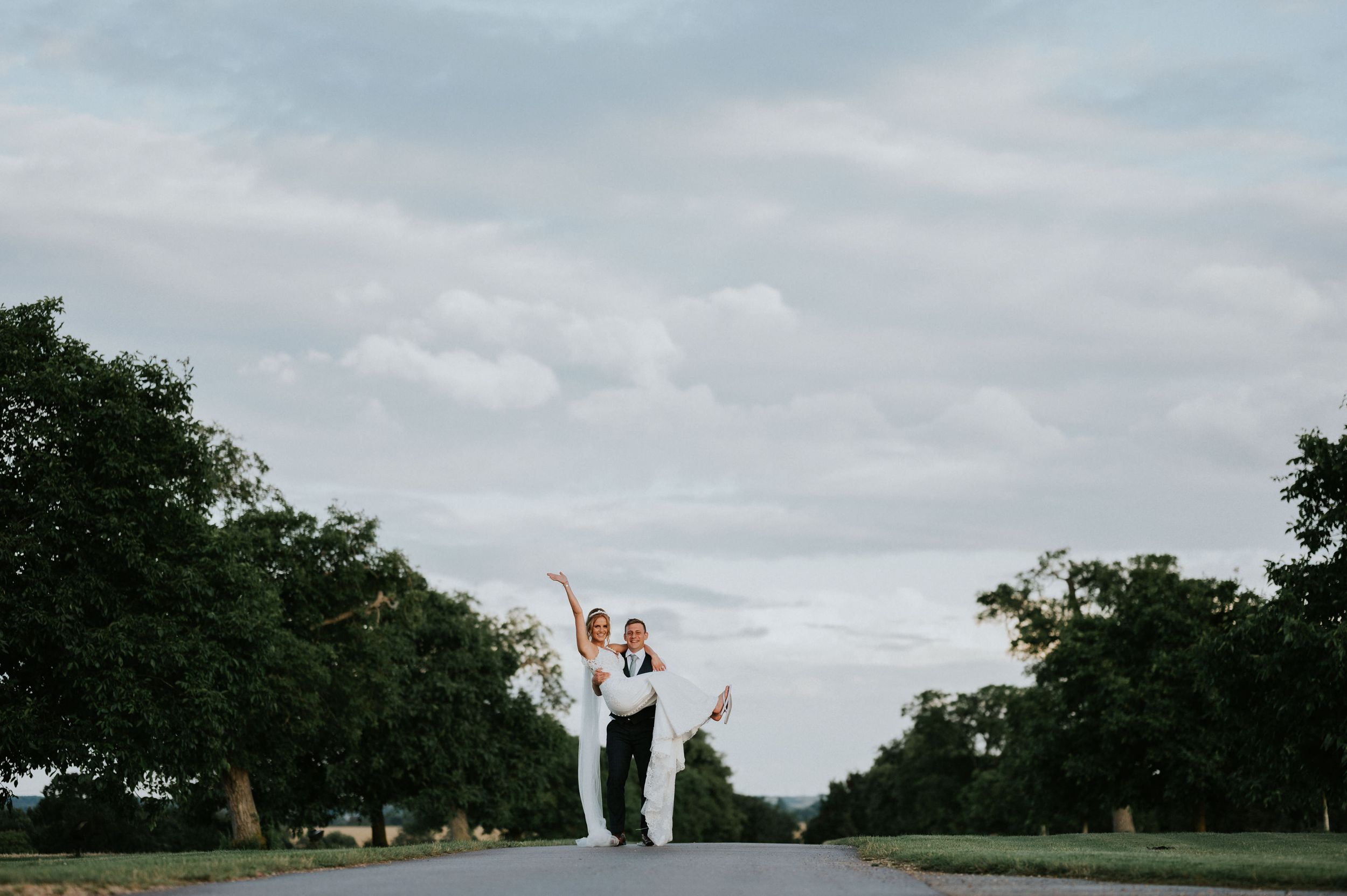Couple celebrating with joyful lift pose on empty road under cloudy sky Bassmead Manor Barns.