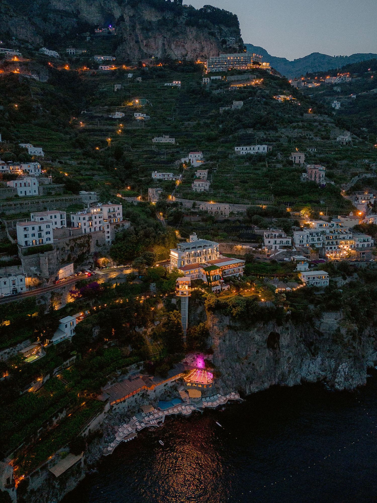 Aerial night view of Hotel Santa Caterina in the Amalfi Coast hillside with buildings cascading down cliffs to the Mediterranean Sea.
