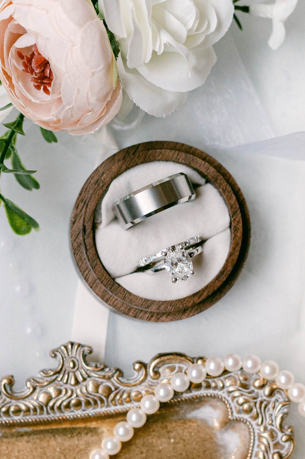 Wedding rings displayed on wooden coasters surrounded by white and blush roses with pearl decorations.