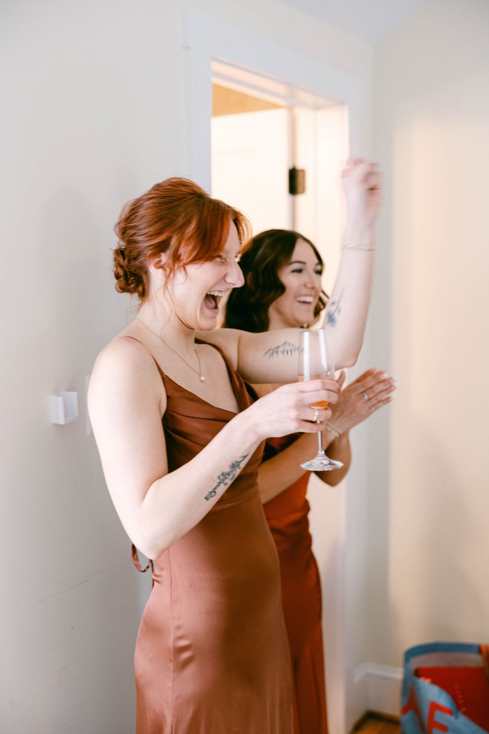 Bridesmaids in rust-colored dresses sharing champagne and laughter during wedding preparations.