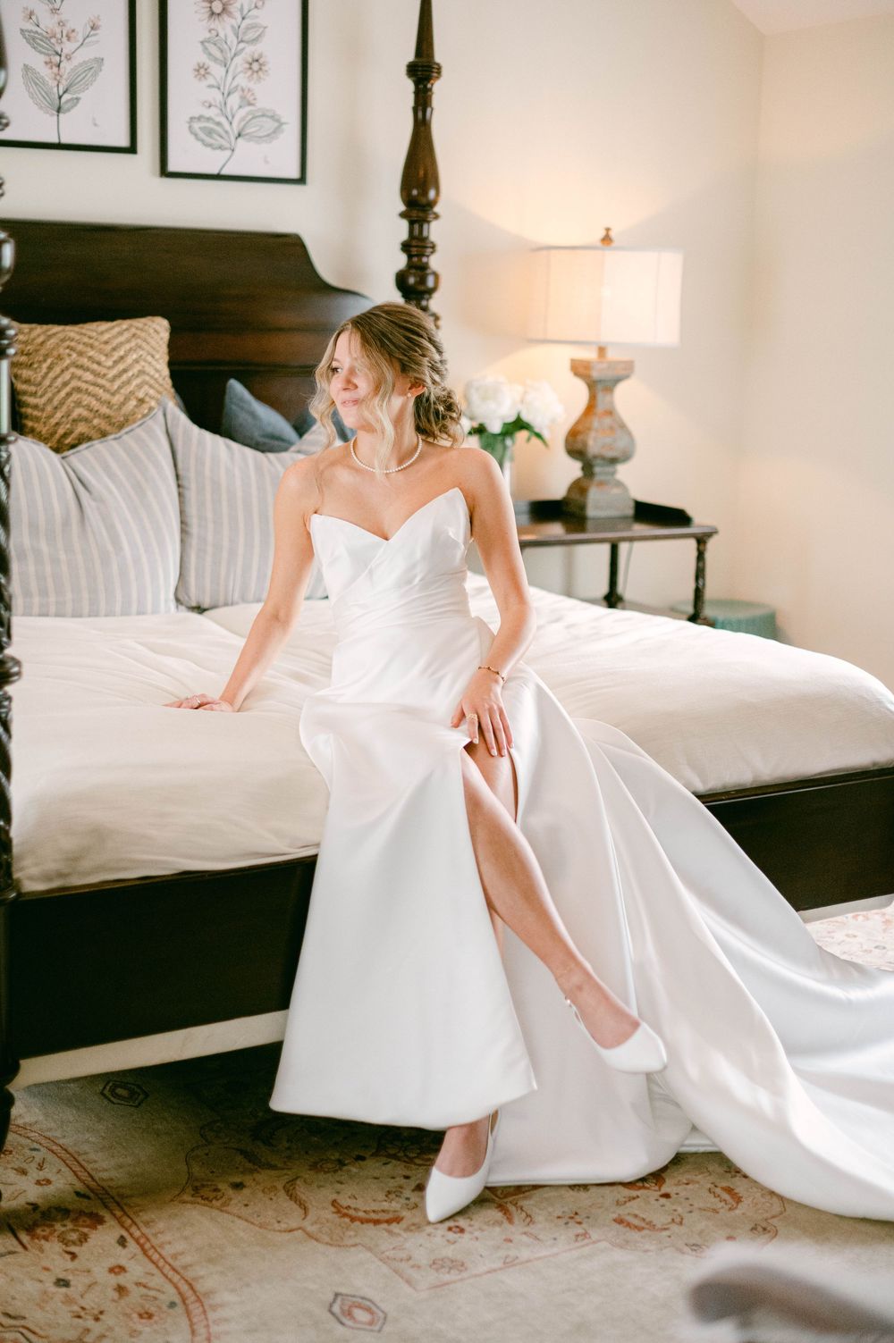 Elegant bride in strapless white wedding gown sitting on edge of four-poster bed.
