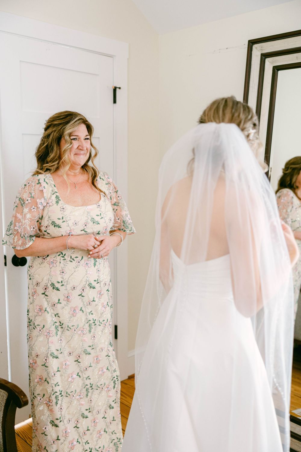 Wedding preparation scene with floral dress and white bridal gown reflected in framed mirror.