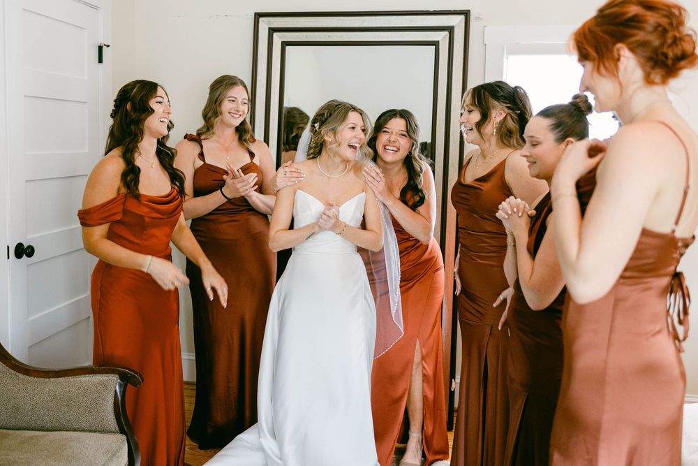 Joyful wedding party scene with bridesmaids in rust-colored dresses surrounding bride in white.