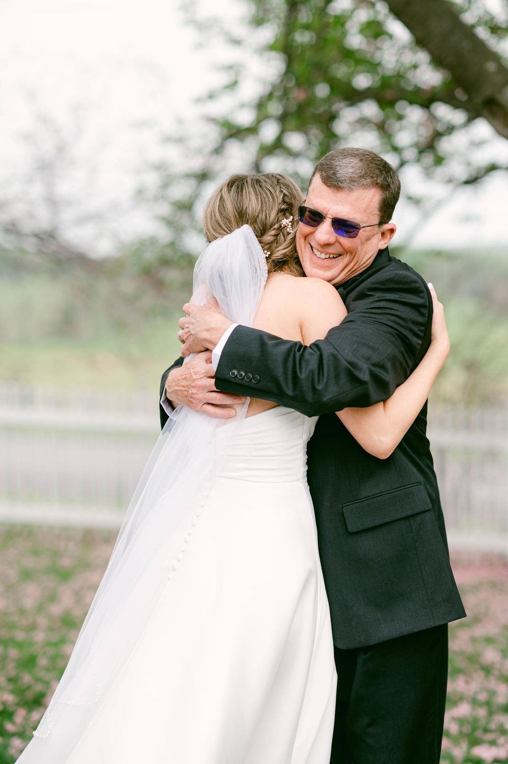 A joyful embrace between newlyweds, with the groom in black suit smiling while hugging his bride in white outdoors.