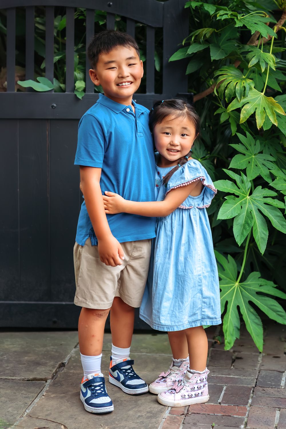 Brother and sister hug and smile next to black fence and pretty green plants during family session at UGA Founders Garden in Athens GA