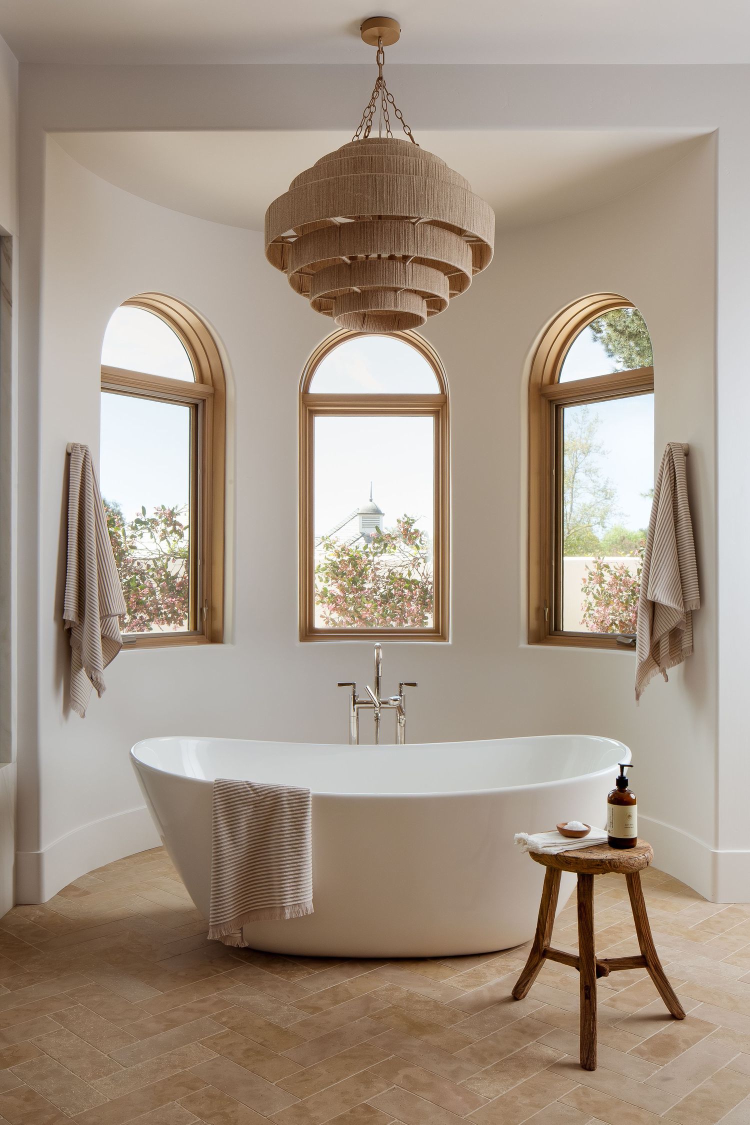 Modern bathroom featuring a freestanding white tub, arched windows, and a tiered woven pendant light in warm natural tones.