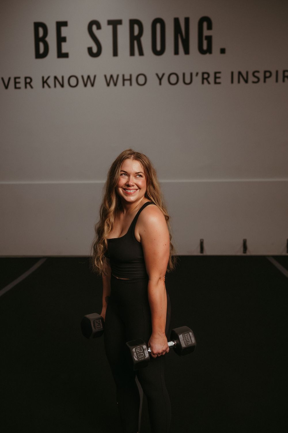 Athletic woman in black workout attire holding dumbbells in a gym with motivational text on the wall.