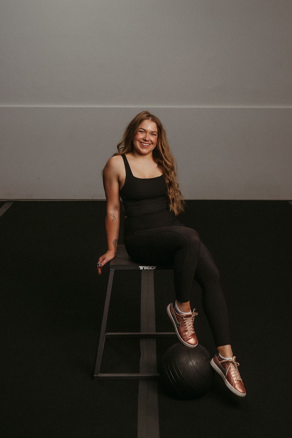 Athletic figure in black workout attire sits on bench with medicine ball in dark studio setting.