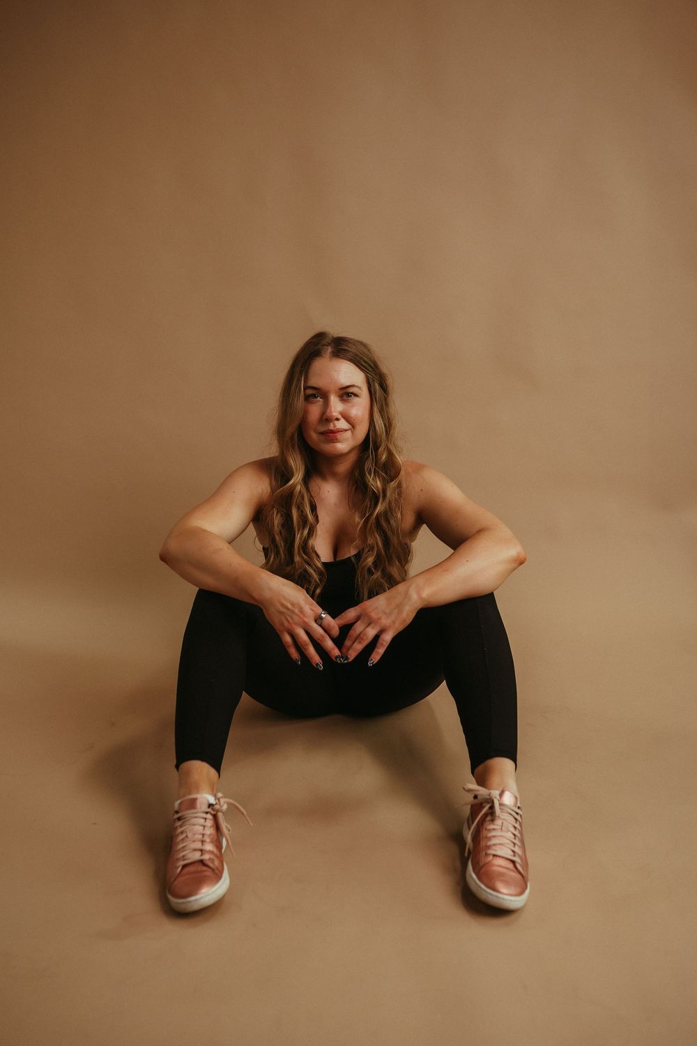 Woman in athletic wear sitting on floor against warm brown backdrop wearing metallic sneakers.