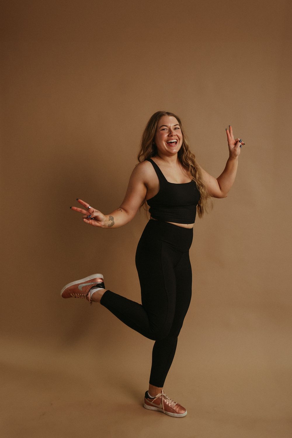 Fitness enthusiast in black workout attire performs dynamic dance poses against warm brown backdrop.